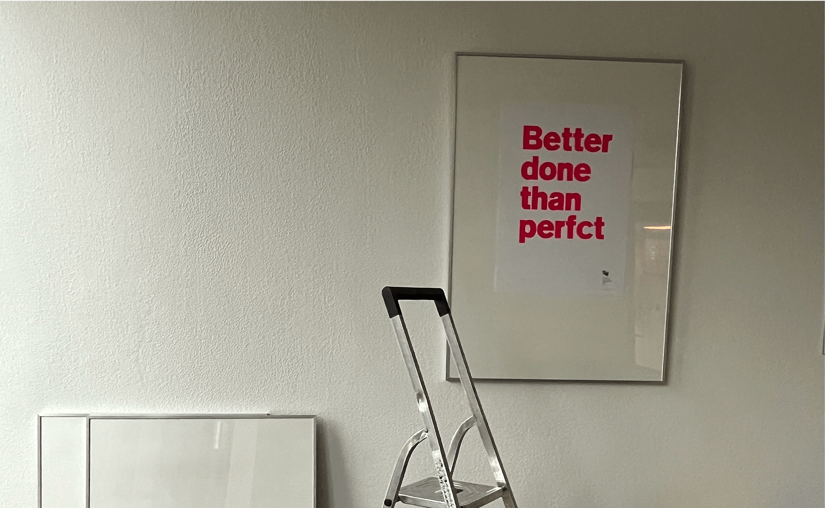 Photo of ladder under framed 'Better done than perfct' poster on white wall