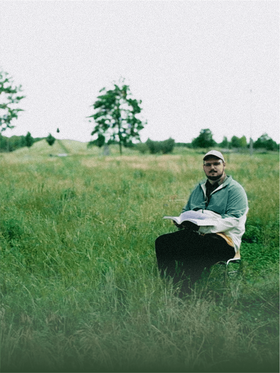 Denis Malinko sitting on a chair reading in a grassy field with trees