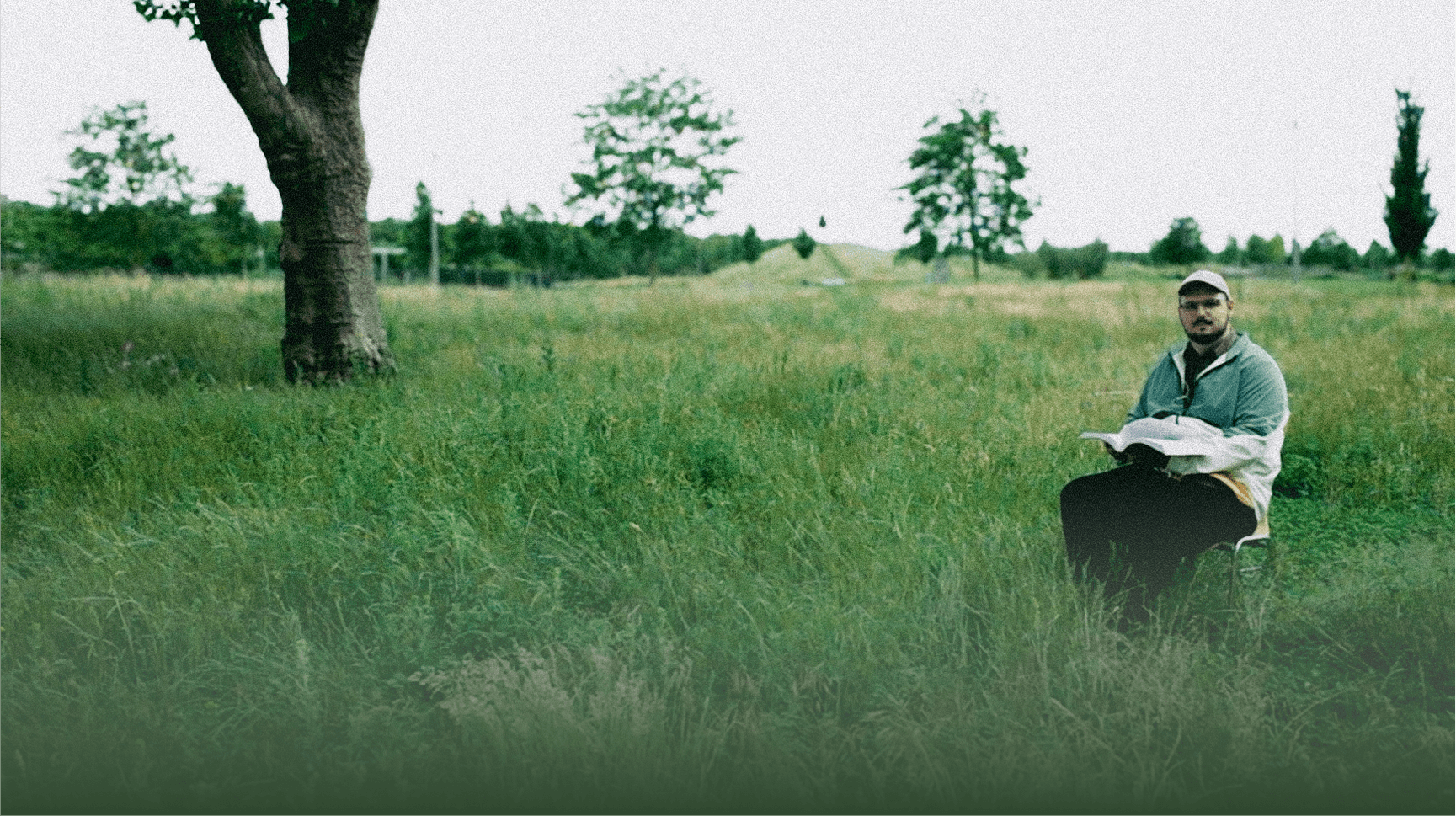 Denis Malinko sitting on a chair reading in a grassy field with trees
