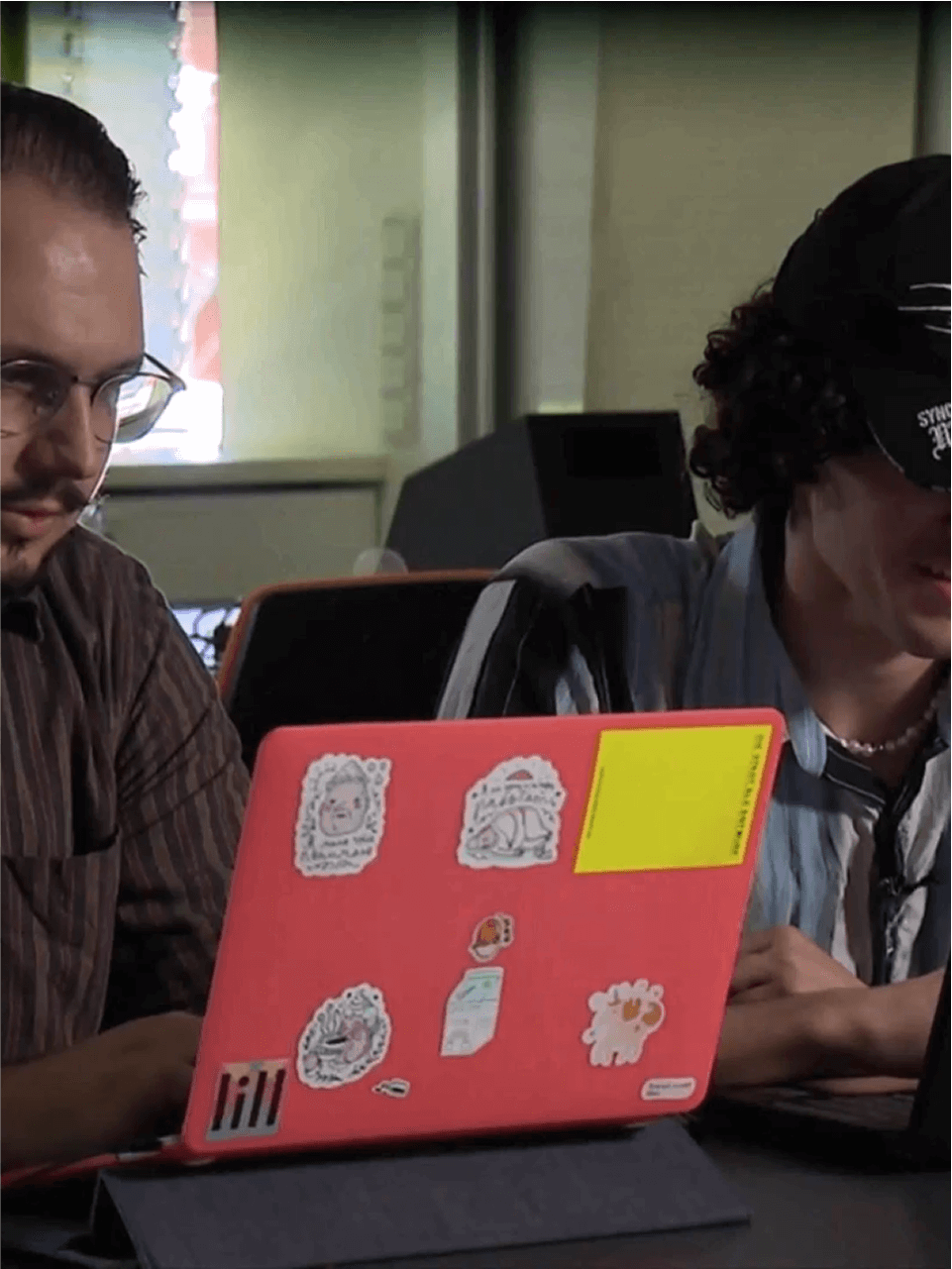 Two students working on laptops in classroom, one with bright sticker-covered case