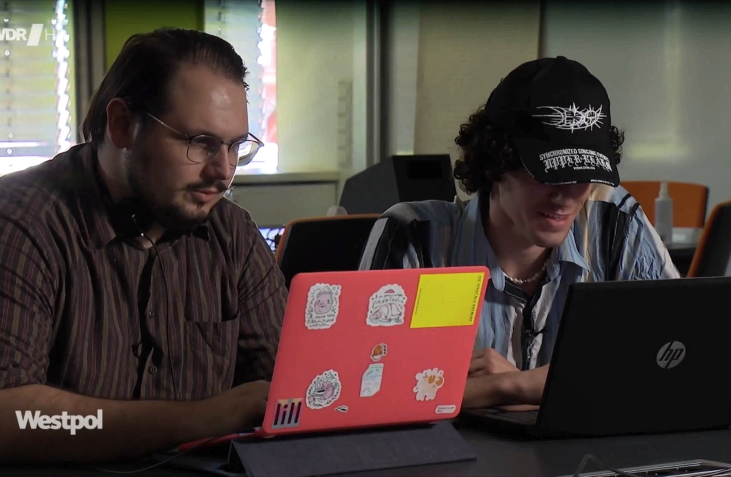 Two students working on laptops in classroom, one with bright sticker-covered case