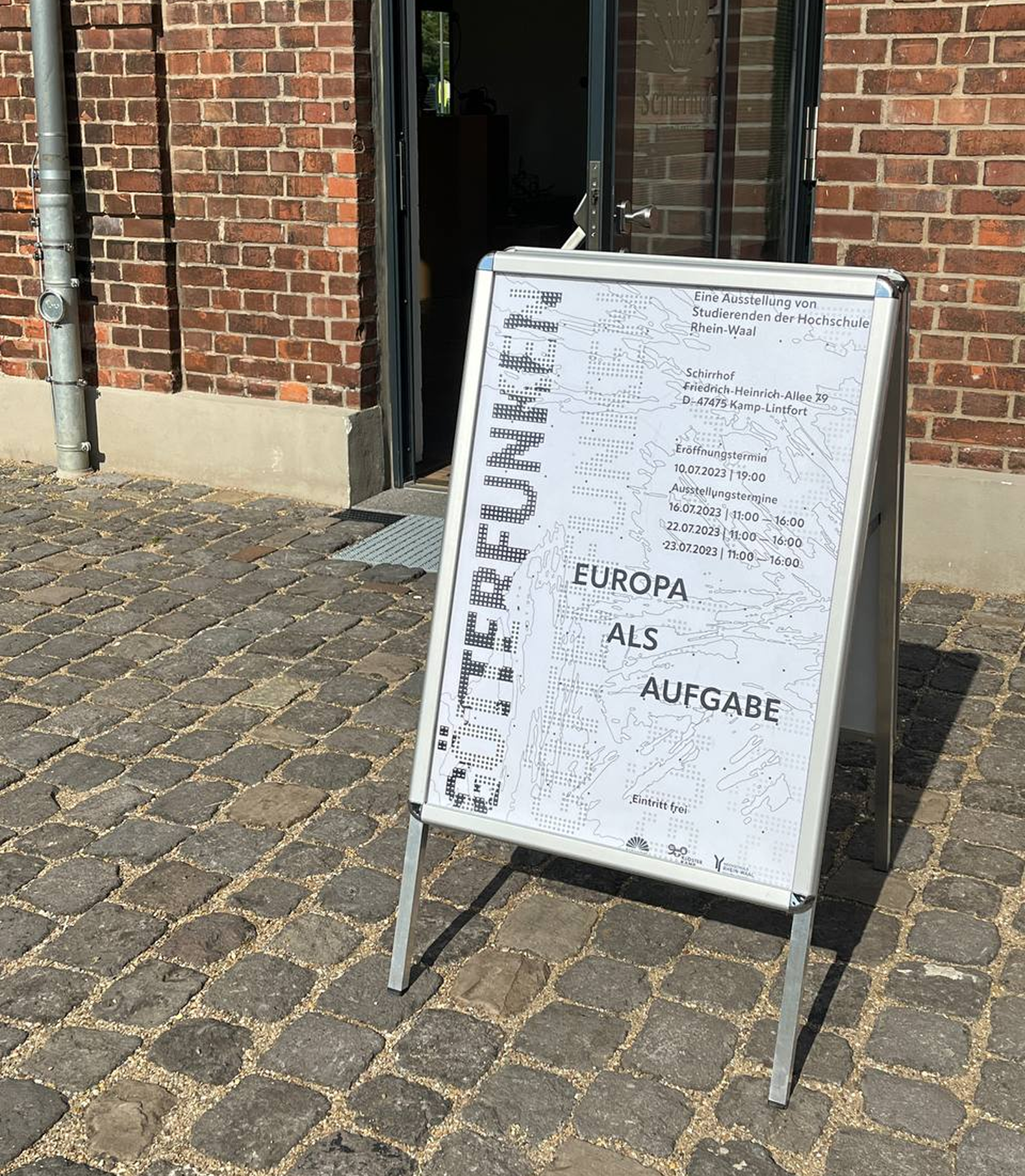 Poster board for GÖTTERFUNKEN exhibition placed outside a brick building