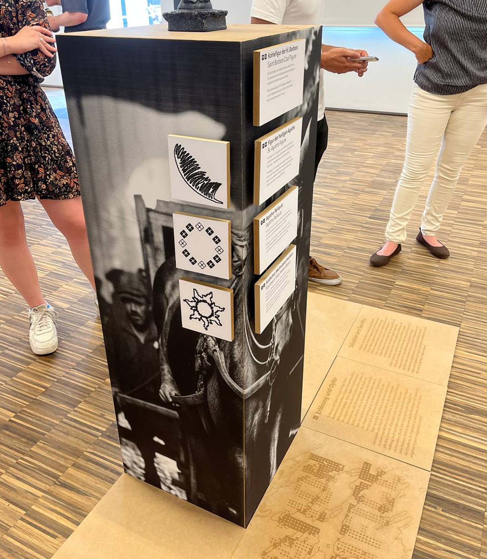 Exhibition pedestal with symbolic icons and printed text, surrounded by visitors